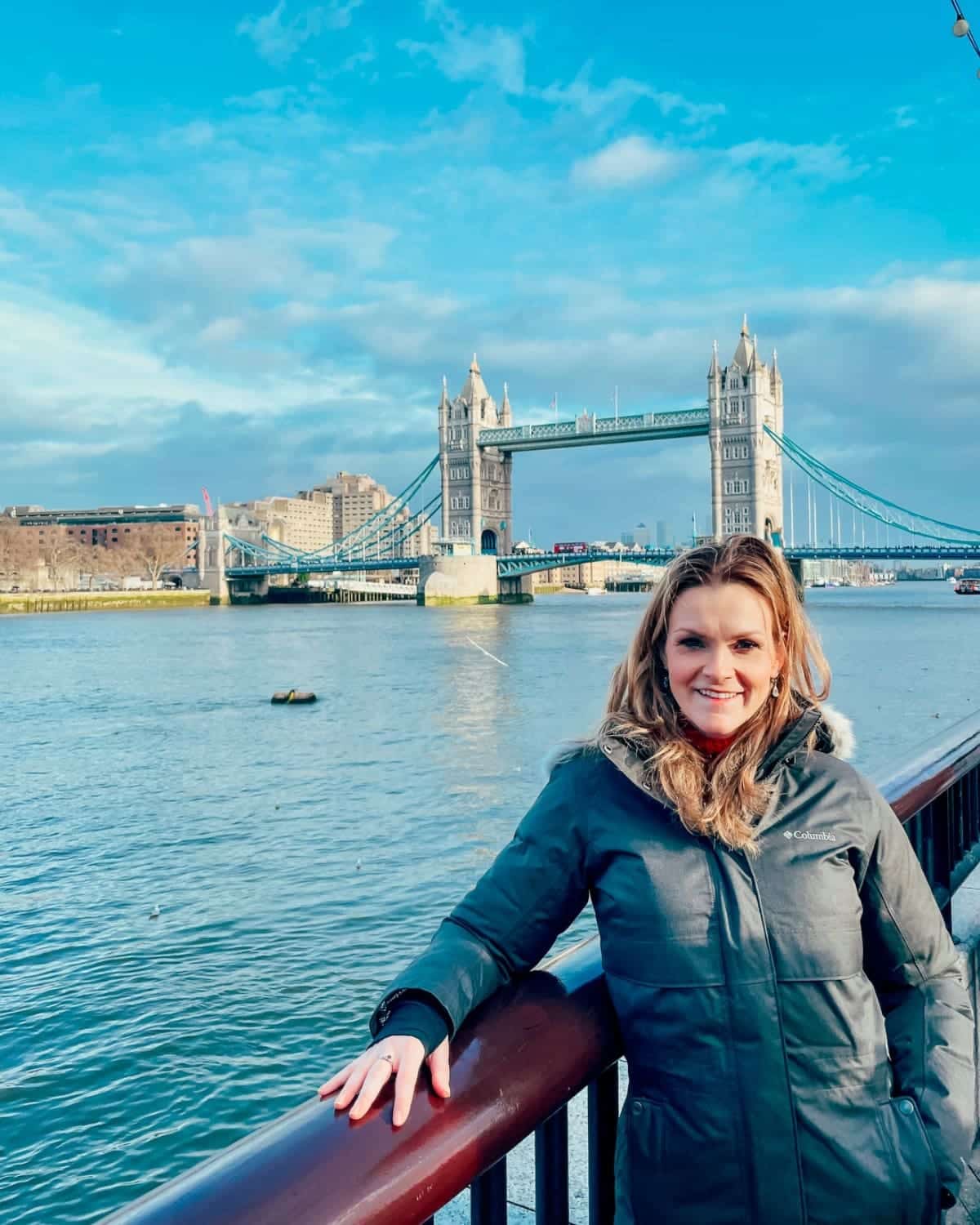 England 11 Michelle L. Jensen (Travel HerStory) smiles beside the River Thames with Tower Bridge in the background in London, England, United Kingdom, wearing a green Columbia winter coat on a sunny, cold day.