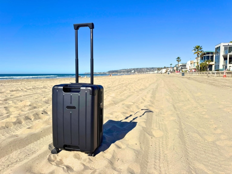 The BlackVoyage AirCabin Pro black carry on suitcase on a sandy beach with it's handle extended under a clear sky.