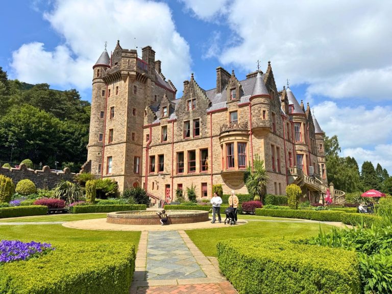 Exterior view of Belfast Castle in Northern Ireland, United Kingdom, from the formal gardens, showcasing the Scottish Baronial-style sandstone facade with conical turrets, slate roofs, and red-trimmed windows, surrounded by manicured box hedges, a central fountain feature, colorful flower beds, and a stone pathway leading to the entrance on a sunny day at Cave Hill, Northern Ireland.