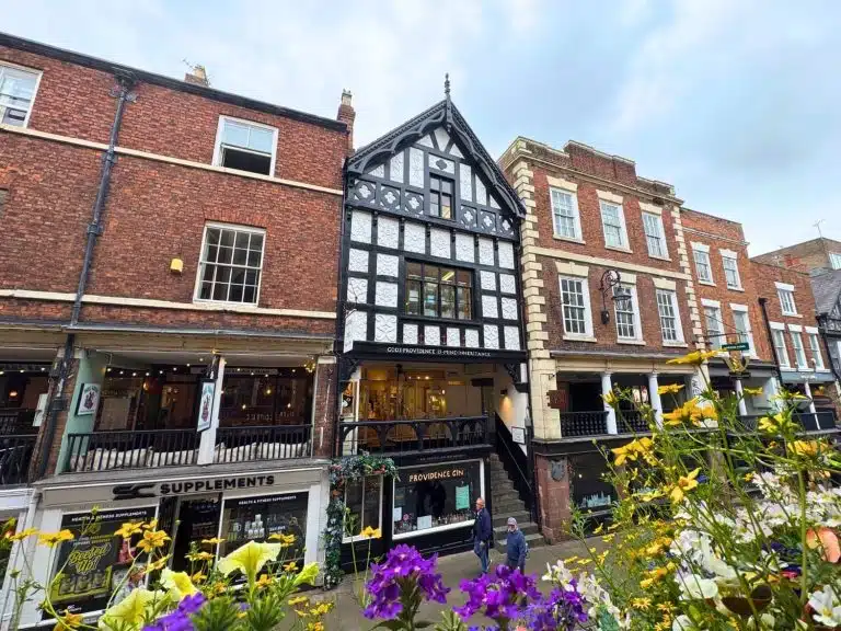 God's Providence House on Watergate Street in Chester, England, United Kingdom, a striking black-and-white half-timbered building dating from the 17th century, bearing the inscription "God's Providence Is Mine Inheritance" on its facade. The Tudor-style structure with ornate carved panels and a decorative gabled roofline stands between red brick Georgian neighbours, with Chester's characteristic two-level Rows visible below. A Providence Gin shop occupies the ground floor, and colorful yellow and purple flowers bloom in the foreground on an overcast day.