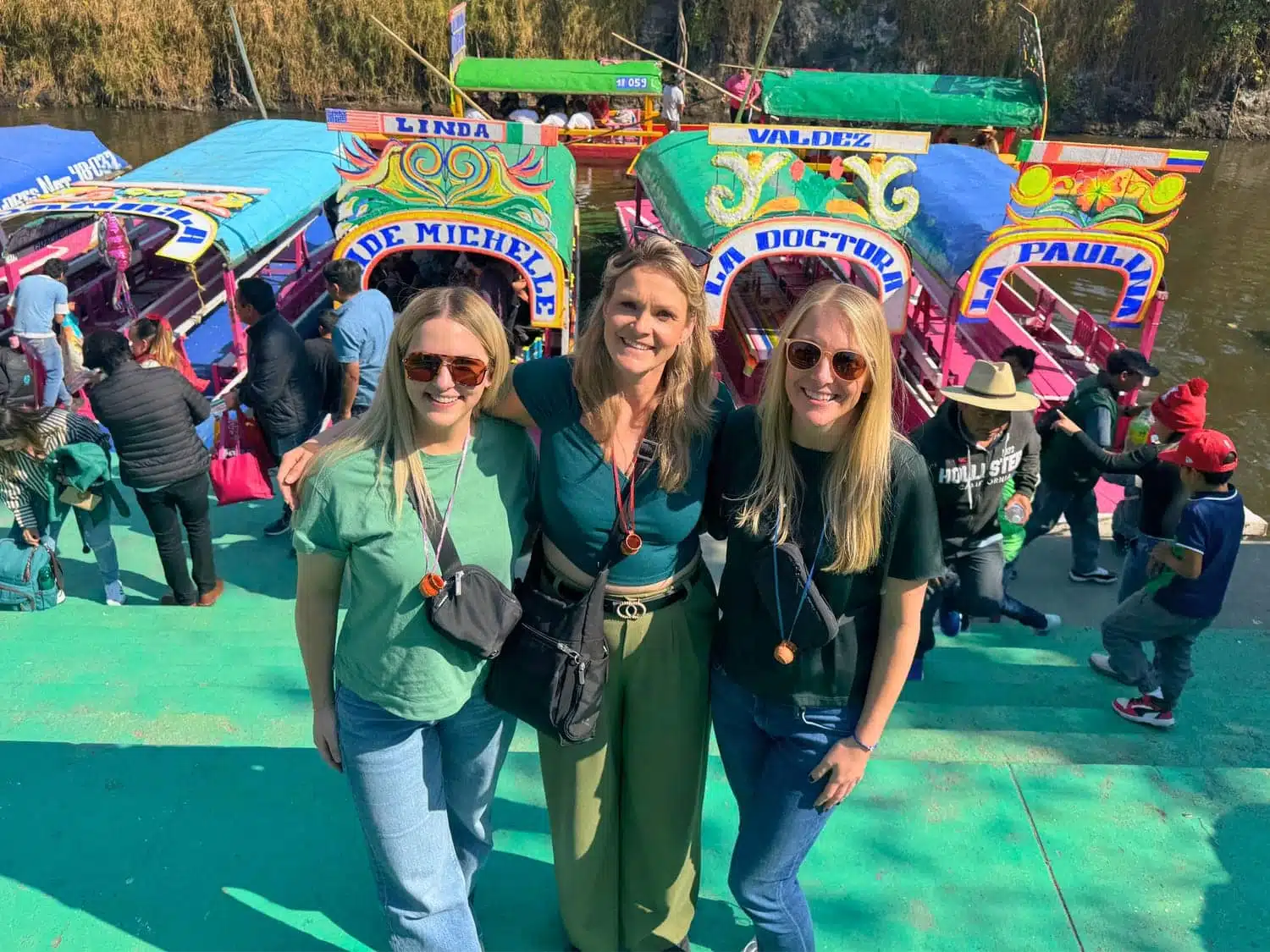 Michelle L. Jensen (Travel HerStory) and friends smiling on a dock at Xochimilco, Mexico City, Mexico, with vibrantly painted and flower-decorated trajinera boats behind them named "De Michelle," "Linda," "Valdez," and "La Doctora," with crowds of visitors boarding nearby.