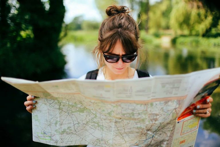 Young brunette woman with hair in a messy bun and Ray-Ban sunglasses reading a large paper road map outdoors, with a blurred green river landscape in the background.