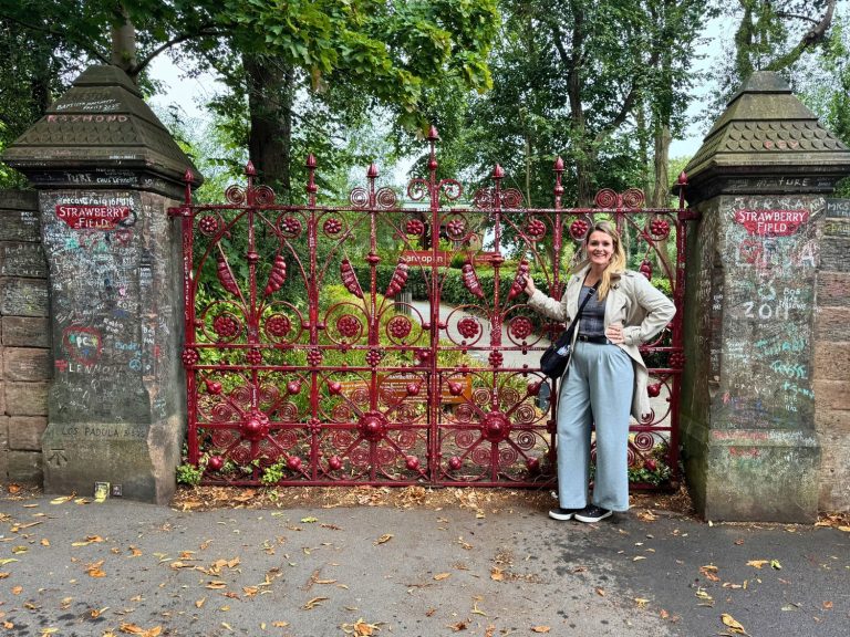 Michelle L. Jensen (Travel HerStory) smiling and posing beside the iconic red ornate iron gates of Strawberry Field in Liverpool, England, United Kingdom, with fan-written graffiti and tributes covering the stone gateposts, autumn leaves on the ground, and lush green trees and gardens visible behind the gates.