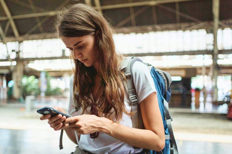 A young woman with long brown hair wearing a white t-shirt and carrying a large blue travel backpack looks down at her iPhone in a decorative case while standing in an open-air transit station with a steel-beam roof.