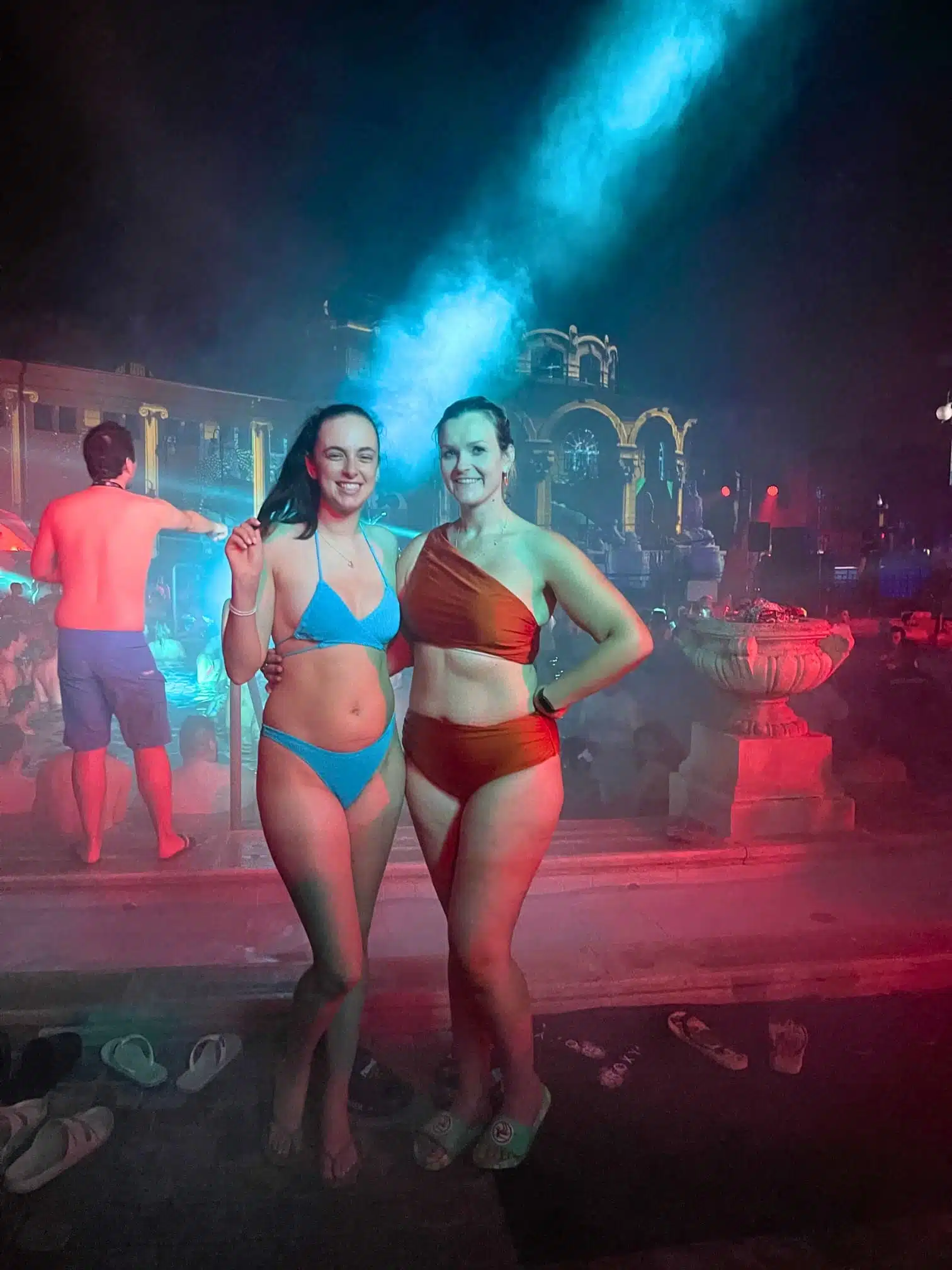 Home 34 Michelle L. Jensen (Travel HerStory) and a friend smiling in bikinis at the SPARTY night pool party at Széchenyi Thermal Bath in Budapest, Hungary, standing poolside with steam rising, blue laser lights, and the illuminated arched architecture of the historic bathhouse glowing in the background amid a crowd of party-goers.