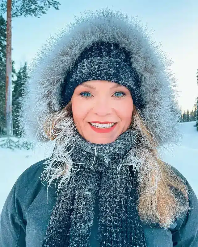 Home 25 Michelle L. Jensen (Travel HerStory) dressed in a winter coat with faux-fur-lined hood, wool hat, and a scarf stands in a snowy landscape at the Ukka-Luosto Fell hike in Finland, with her hair coated in ice crystals.
