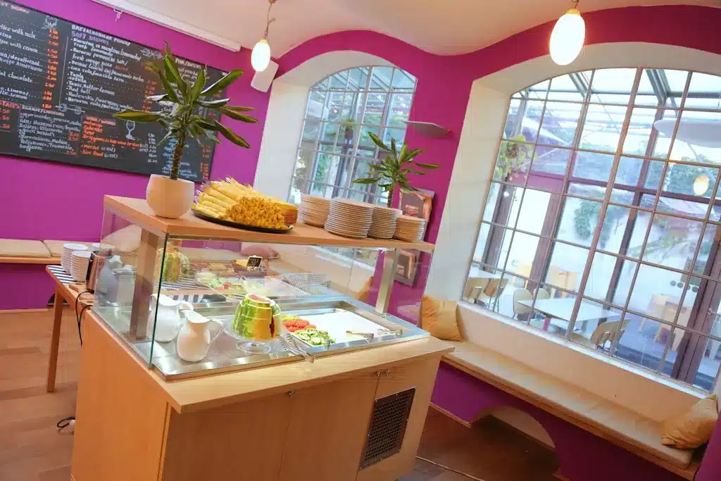 Best Places to Stay in Ljubljana, Slovenia: Key Areas & Top-Rated Hotels & Hostels 30 Bright breakfast buffet area at Hostel Celica in Metelkova, Ljubljana, Slovenia. A glass-fronted display case holds fresh fruit including watermelon and cucumber, topped with sliced pineapple. Hot-pink walls, arched barred windows, a chalkboard menu, and a window bench seat complete the space.