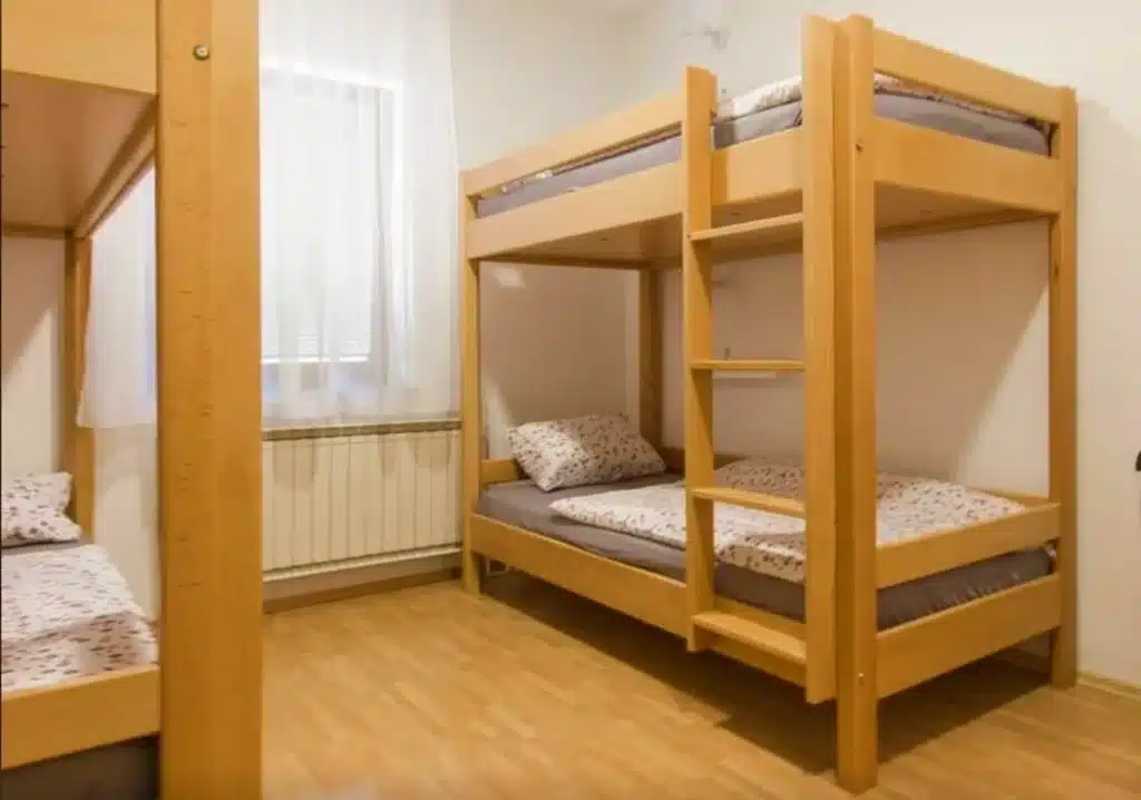 Best Places to Stay in Ljubljana, Slovenia: Key Areas & Top-Rated Hotels & Hostels 32 Shared dormitory room at Hostel Vrba in Ljubljana, Slovenia, featuring light pine wood bunk beds with gray mattresses and floral pillowcases, light wood laminate flooring, white walls, a radiator, and a sheer-curtained window providing natural light.