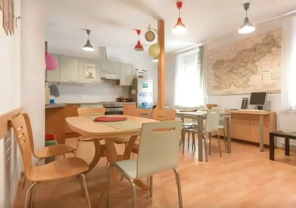 Best Places to Stay in Ljubljana, Slovenia: Key Areas & Top-Rated Hotels & Hostels 31 Common room and shared kitchen at Hostel Vrba in Ljubljana, Slovenia. The bright space features light wood dining tables and mixed chairs, colorful pendant lights, a large wall map of Slovenia, a shared kitchen with refrigerator and stove, a desktop computer workstation, and wood laminate floors.