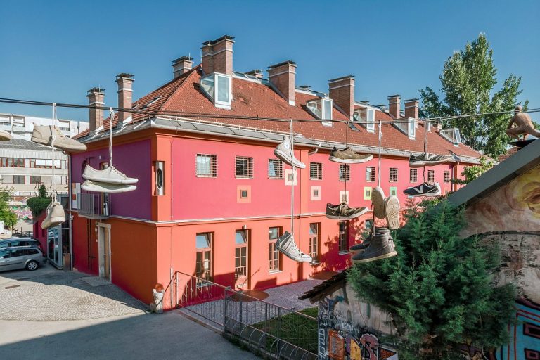 Exterior of Hostel Celica at Metelkova Mesto in Ljubljana, Slovenia, a former military prison converted into a boutique hostel. The vivid coral-pink and magenta building with a red-tiled mansard roof has barred windows, and oversized shoes hang from wires above the courtyard as an art installation.