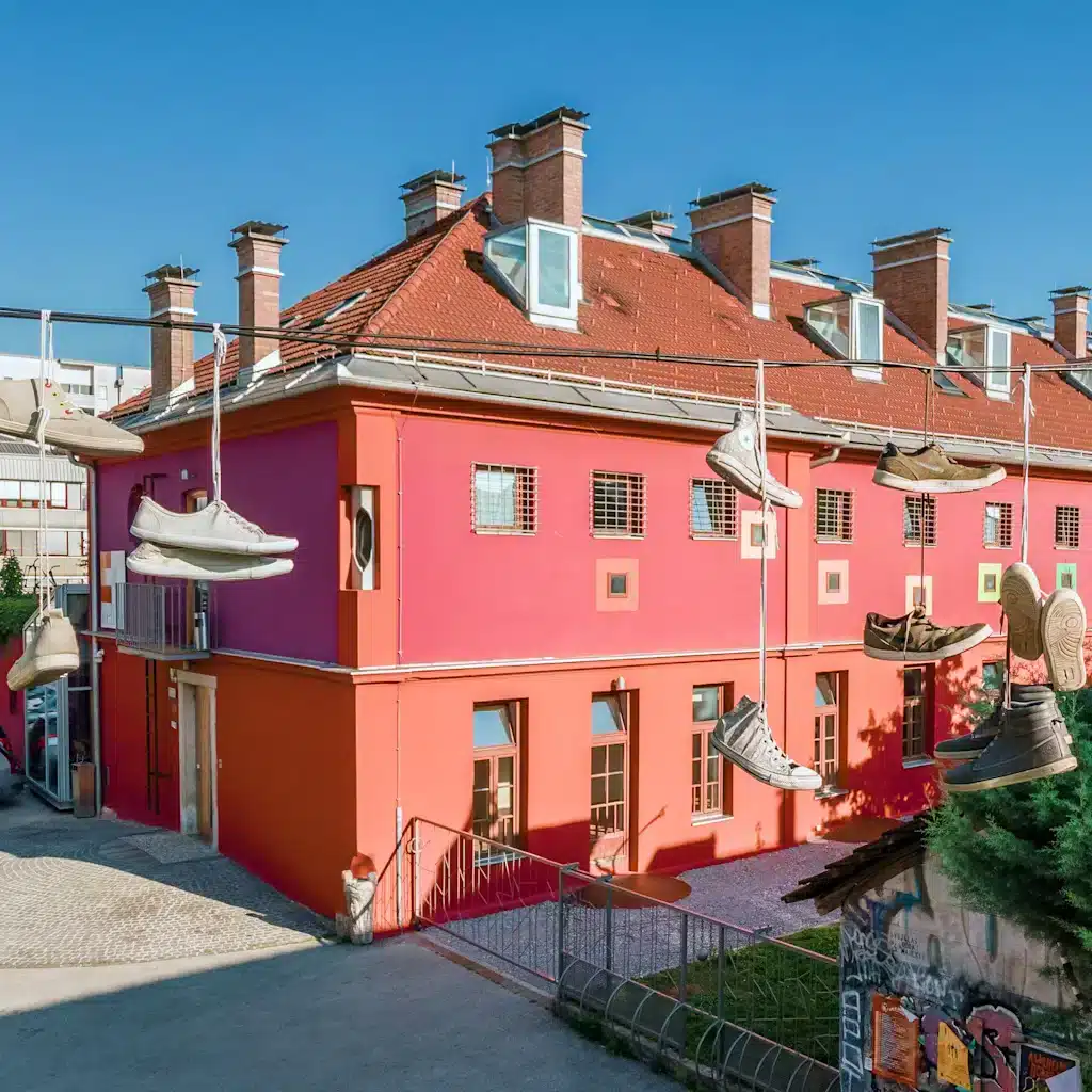 Best Places to Stay in Ljubljana, Slovenia: Key Areas & Top-Rated Hotels & Hostels 29 Exterior of Hostel Celica at Metelkova Mesto in Ljubljana, Slovenia, a former military prison converted into a boutique hostel. The vivid coral-pink and magenta building with a red-tiled mansard roof has barred windows, and oversized shoes hang from wires above the courtyard as an art installation.