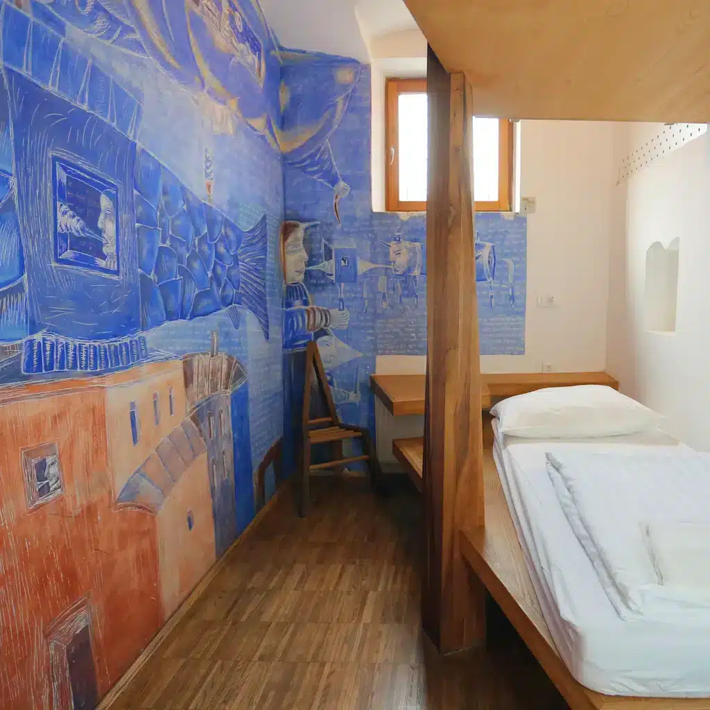 Best Places to Stay in Ljubljana, Slovenia: Key Areas & Top-Rated Hotels & Hostels 10 Interior of an artist-designed room at Hostel Celica in Ljubljana, Slovenia, a converted former prison. The narrow room features a vivid hand-painted blue-and-terracotta surrealist mural covering the walls, a wooden single bed with white linens, wood-plank ceiling, and small barred window.