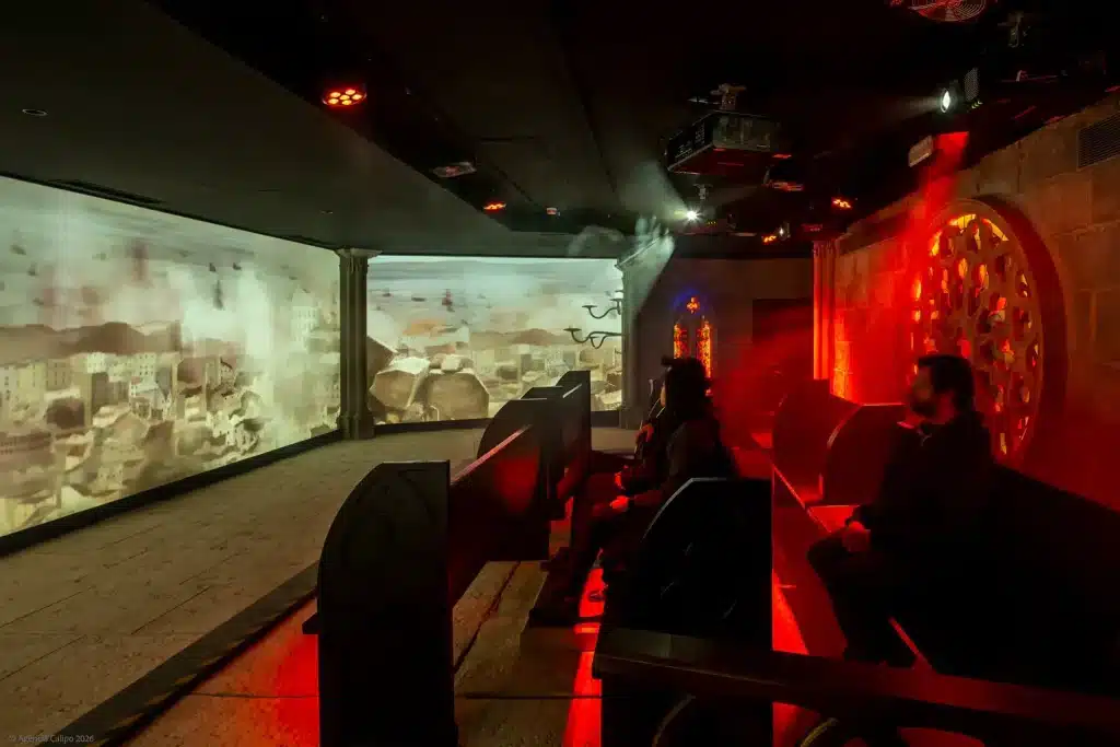 22 of the Best Things to Do in Lisbon, Portugal (My Budget-Friendly Picks) 24 Two visitors seated in a darkened immersive theater room at the Lisbon Story Centre, watching a dramatic projected animation depicting the 1755 Lisbon earthquake and destruction across multiple large screens, with red atmospheric lighting and stone arch décor surrounding them.