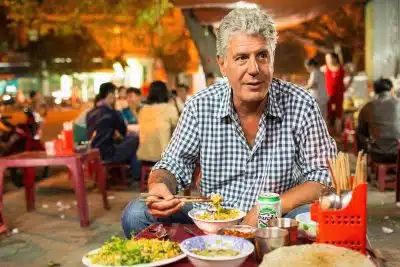 12 Travel TV Shows to Watch for Inspiration + Where to Stream Each Series 2 Anthony Bourdain, TV host in a blue gingham shirt, eating with chopsticks at a low plastic table on a bustling Vietnamese street food stall at night, with bowls of rice and noodle dishes, a Huda beer can, and blurred diners in warm ambient lighting behind him while filming his travel TV series Parts Unknown.