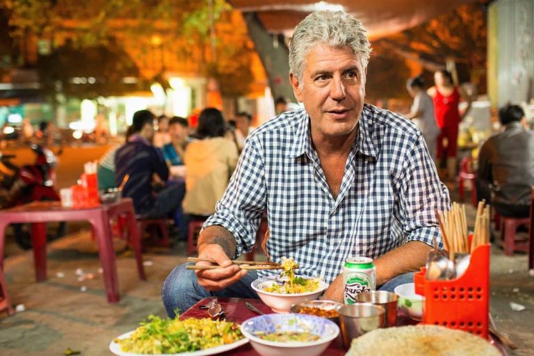 Anthony Bourdain, TV host in a blue gingham shirt, eating with chopsticks at a low plastic table on a bustling Vietnamese street food stall at night, with bowls of rice and noodle dishes, a Huda beer can, and blurred diners in warm ambient lighting behind him while filming his travel TV series Parts Unknown.