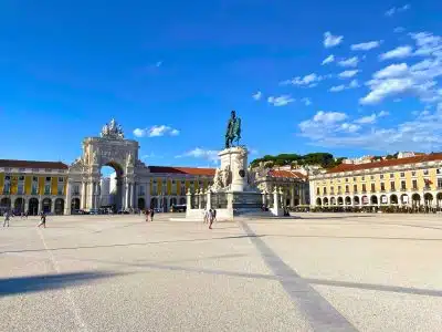 2-Day Lisbon Itinerary: Budget-Friendly Plan for a Weekend in Portugal's Capital 3 Praça do Comércio in Lisbon, Portugal, featuring the ornate white Arco da Rua Augusta triumphal arch, the bronze equestrian statue of King José I on a tall marble pedestal, and surrounding yellow neoclassical government buildings under a vivid blue sky with scattered clouds.