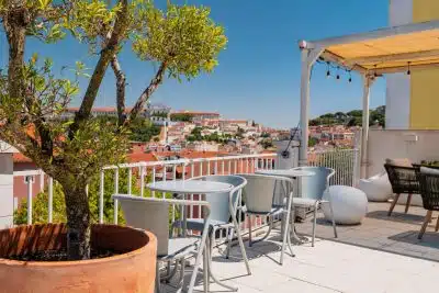 Where to Stay in Lisbon, Portugal: Best Areas + Top-Rated Hotels & Hostels 5 Quinta Colina by Shiadu rooftop terrace in Lisbon, Portugal, with light blue outdoor seating, round tables, and a large terracotta potted tree in the foreground, overlooking a panoramic view of Lisbon's terracotta rooftops and the Graça hilltop with Igreja da Graça visible under a clear blue sky.