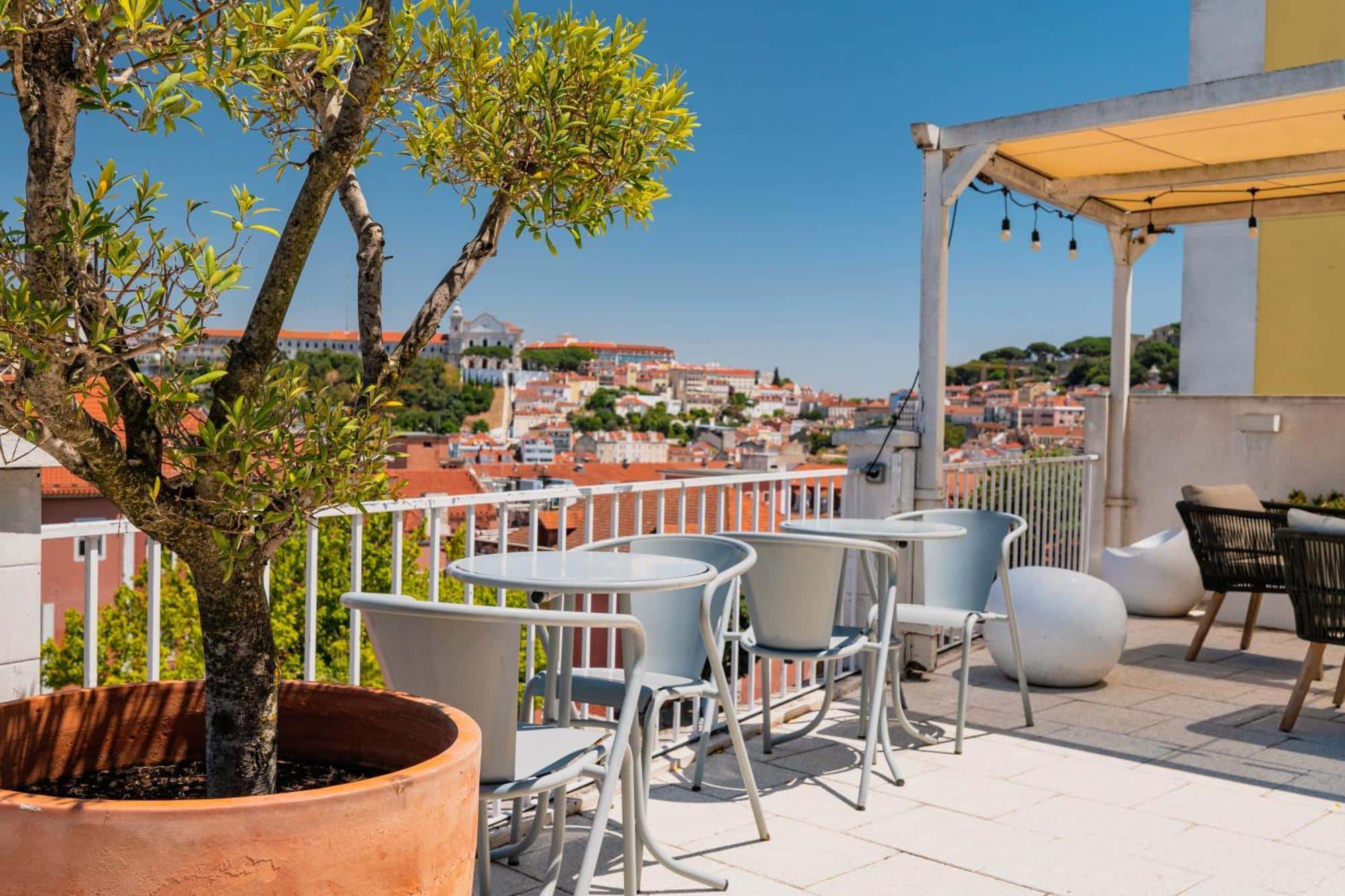 Quinta Colina by Shiadu rooftop terrace in Lisbon, Portugal, with light blue outdoor seating, round tables, and a large terracotta potted tree in the foreground, overlooking a panoramic view of Lisbon's terracotta rooftops and the Graça hilltop with Igreja da Graça visible under a clear blue sky.