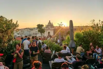 Is Lisbon Safe for Solo Female Travelers? Top Tips to Keep You Safe 6 Crowded rooftop terrace at Park Bar in Lisbon, Portugal, at golden hour sunset, with guests socializing, holding cocktails, and seated on wooden lounge chairs among tropical palms and lush greenery, overlooking the city skyline and a domed historic church bathed in warm orange light.
