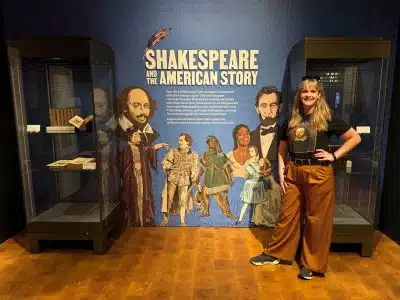 Folger Shakespeare Library Review: Is the Renovated Exhibition Worth a Visit? 1 Michelle L. Jensen (Travel HerStory) poses beside the 'Shakespeare and the American Story' exhibit wall at the Folger Shakespeare Library in Washington, D.C., wearing a William Shakespeare portrait t-shirt, flanked by glass display cases and a colorful mural of historical figures.