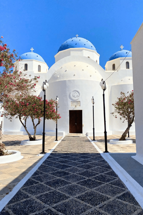 The Church of the Holy Cross in Santorini, Greece, featuring a large white facade and a bright blue dome topped with a cross. The church stands out against a clear sky, as a stone path leads to it with pink flowers blooming to the sides.