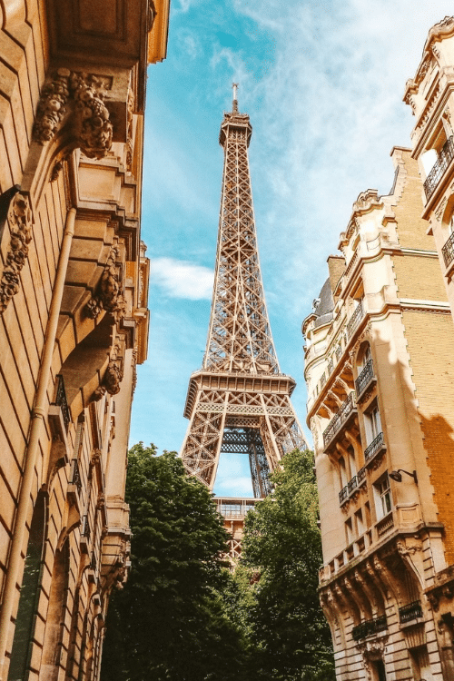View of the Eiffel Tower in Paris, France, from a side street framed by trees, Parisian buildings, and a blue sky.