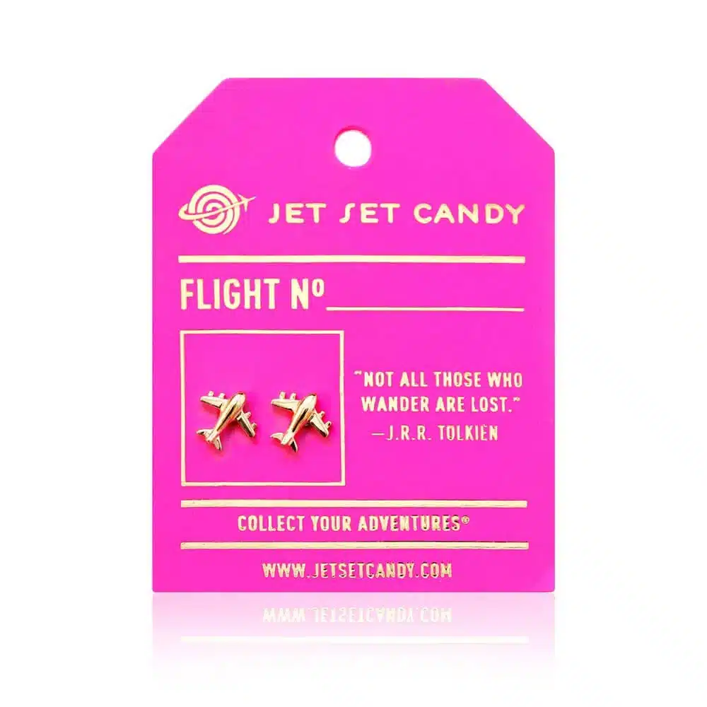 35 Gift Ideas for Someone Traveling to Europe That They'll Actually Use (2026 Guide) 3 Gold airplane stud earrings by Jet Set Candy mounted on a hot pink luggage-tag-style card with gold foil lettering, a blank Flight No line, the J.R.R. Tolkien quote Not All Those Who Wander Are Lost, and the Collect Your Adventures tagline, shot on a white background with a soft reflection.