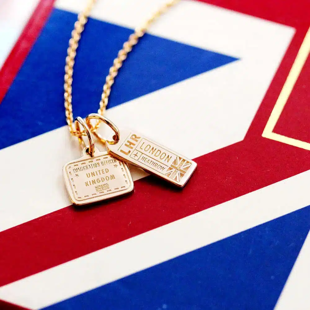 35 Gift Ideas for Someone Traveling to Europe That They'll Actually Use (2026 Guide) 4 Two gold Jet Set Candy travel charms on a gold chain necklace resting on a red, white, and blue Union Jack flag: an LHR London Heathrow luggage-tag charm with a Union Jack detail and a square United Kingdom immigration stamp charm with a dotted passport-stamp border, in a close-up product shot.