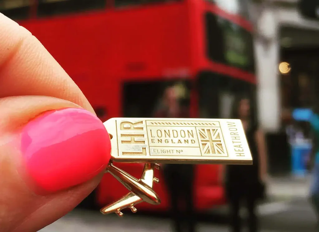 35 Gift Ideas for Someone Traveling to Europe That They'll Actually Use (2026 Guide) 1 Close-up of a hand with hot pink nail polish holding a gold Jet Set Candy LHR London England Heathrow luggage-tag charm engraved with a Union Jack and Flight No line, with a gold airplane charm below it, in front of a blurred red double-decker bus on a street in London, England, United Kingdom.