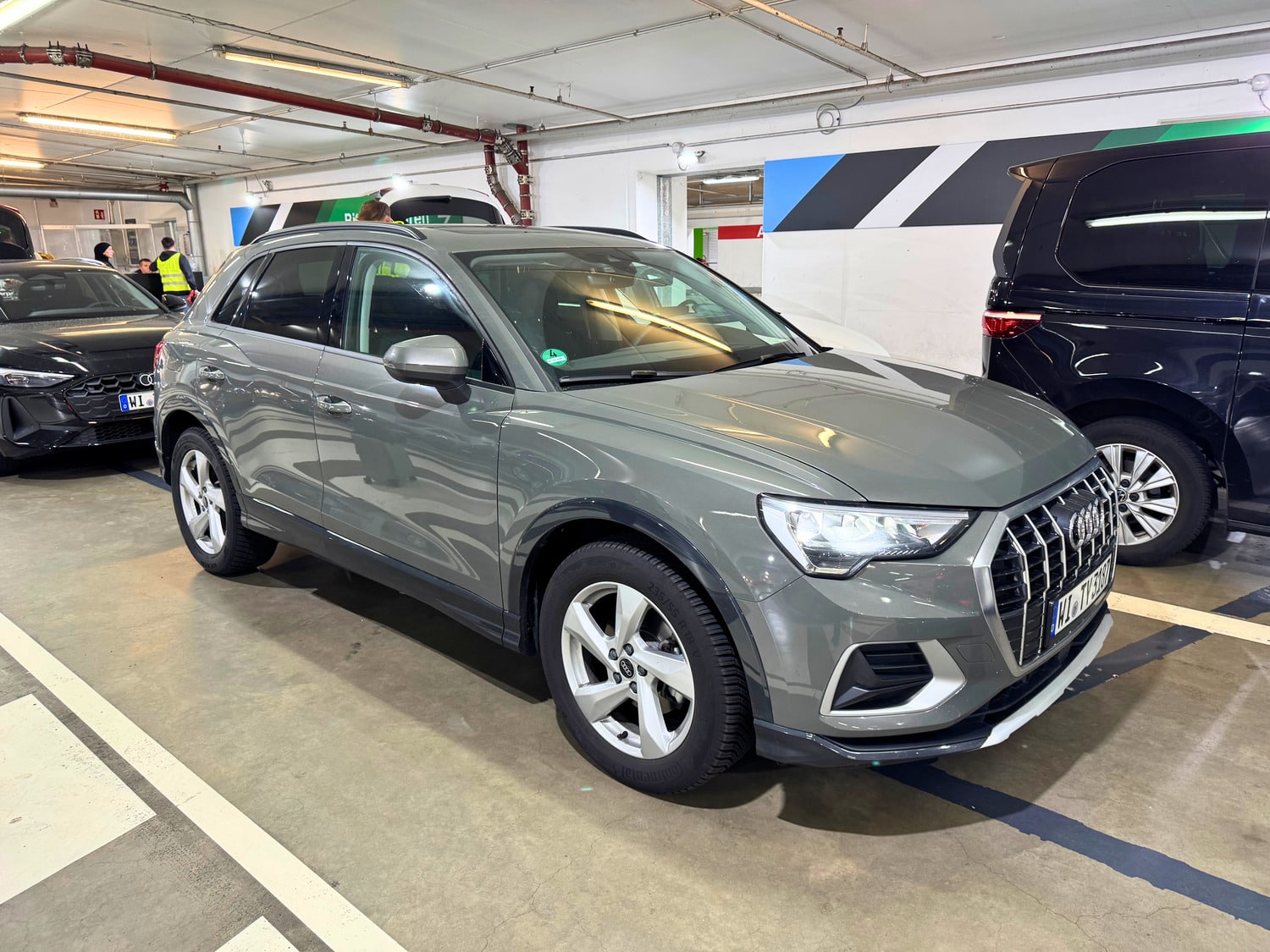 Gray Audi Q3 SUV with German license plates parked in a numbered bay of the rental car return garage at Frankfurt Airport, beside a black Audi sedan and a dark minivan, with a worker in a yellow safety vest and blue, green and black striped walls behind it. Frankfurt am Main, Hesse, Germany.
